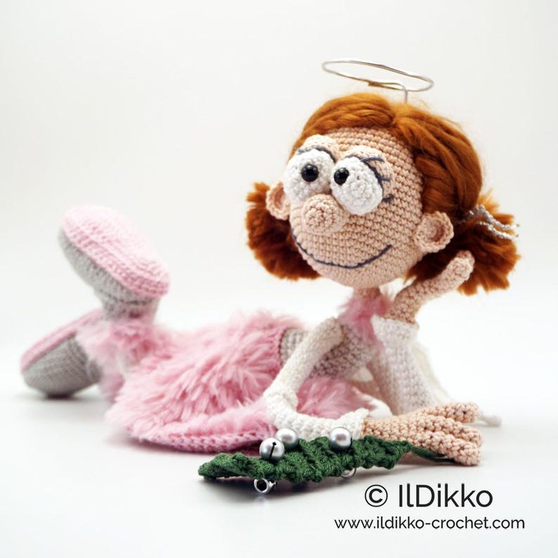May include: A crocheted doll with auburn hair, a silver wire halo, and large white eyes. The doll wears a pink fuzzy skirt, white long sleeves, and grey shoes, while resting on one arm next to a small green crocheted branch with silver bells.