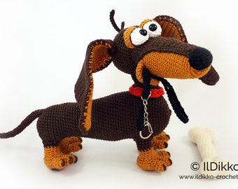 May include: Crocheted dachshund dog with brown and tan fur, large eyes, and a red collar. A black leash and a white bone are next to the dog. The dog is handmade and has a whimsical design. The website address is visible at the bottom.