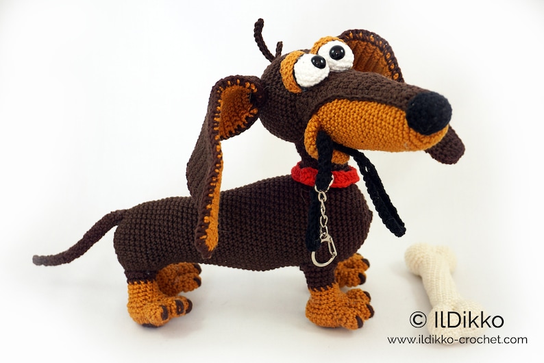 May include: Crocheted dachshund dog with brown and tan fur, large eyes, and a red collar. A black leash and a white bone are next to the dog. The dog is handmade and has a whimsical design. The website address is visible at the bottom.