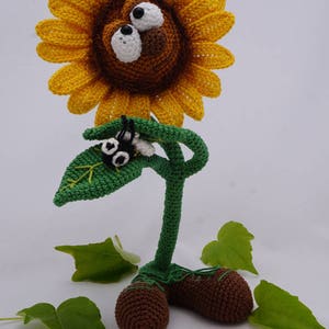 May include: A crocheted sunflower with a brown face and large white eyes. The sunflower has a green stem and leaves, and is standing on two brown crocheted feet. The sunflower is surrounded by green leaves.