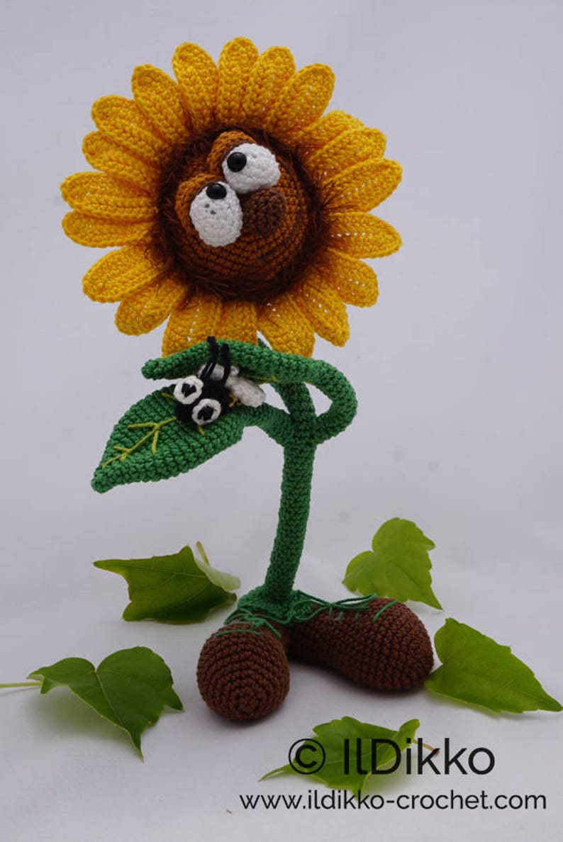 May include: A crocheted sunflower with a brown face and large white eyes. The sunflower has a green stem and leaves, and is standing on two brown crocheted feet. The sunflower is surrounded by green leaves.