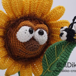 May include: A crocheted sunflower with a brown face and large white eyes. A crocheted bee with black and yellow stripes is perched on a green leaf.