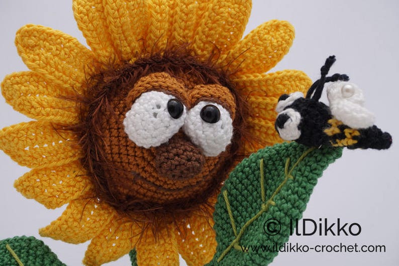 May include: A crocheted sunflower with a brown face and large white eyes. A crocheted bee with black and yellow stripes is perched on a green leaf.