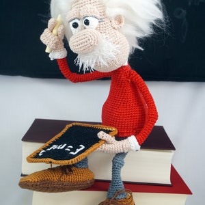 Puede incluir: Mu&ntilde;eco de ganchillo hecho a mano de una figura de Einstein de dibujos animados, con un su&eacute;ter rojo, pantalones grises y zapatos marrones. El mu&ntilde;eco sostiene una peque&ntilde;a pizarra con ecuaciones. La figura tiene pelo blanco y barba blanca. El mu&ntilde;eco est&aacute; sentado encima de una pila de libros.
