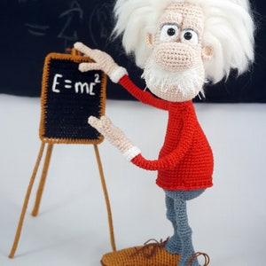 May include: A crocheted doll of a scientist with wild white hair, a white beard, a red long-sleeved shirt, and gray pants. The doll stands next to a small black chalkboard on a wooden easel with the white text E=mc2 written on the board.