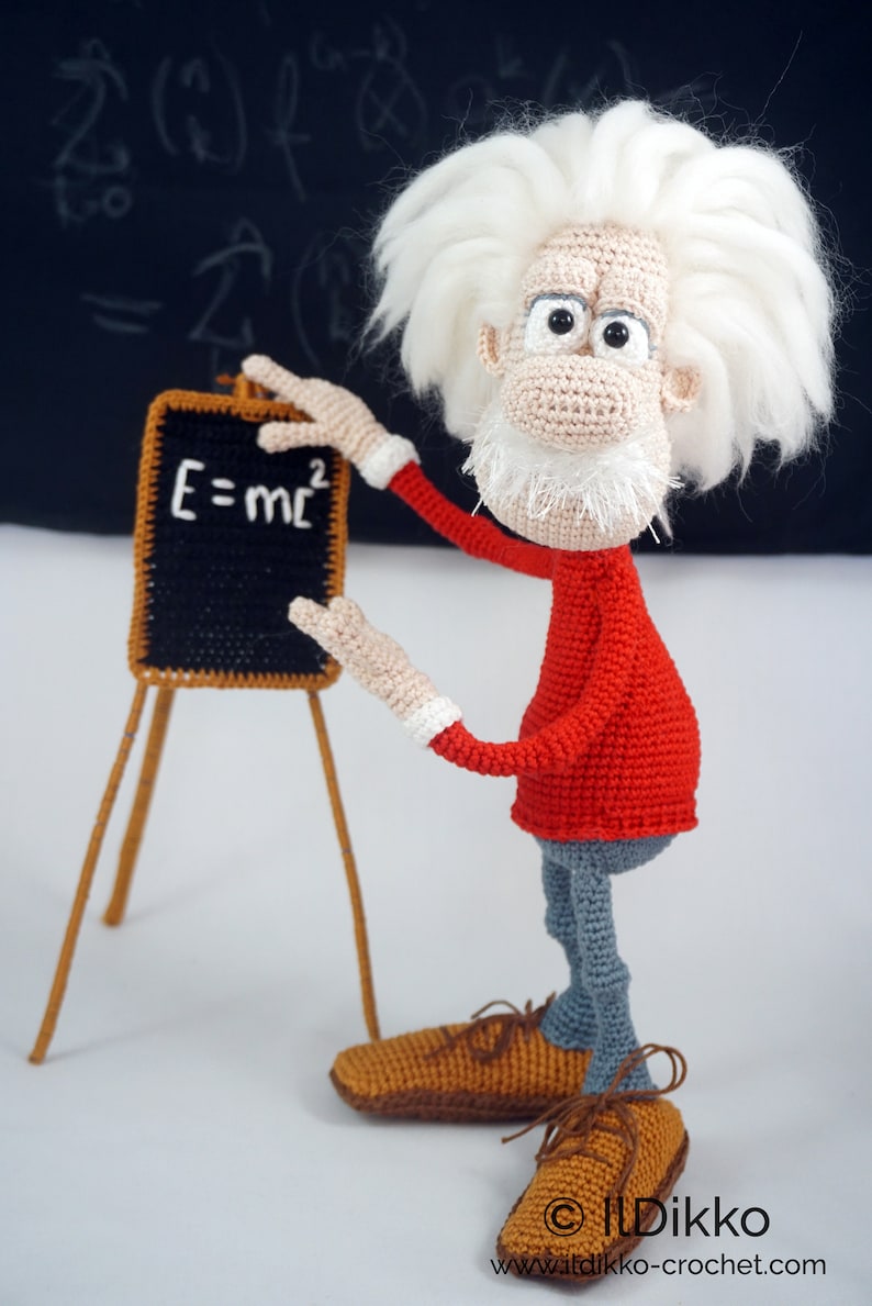 May include: A crocheted doll of a scientist with wild white hair, a white beard, a red long-sleeved shirt, and gray pants. The doll stands next to a small black chalkboard on a wooden easel with the white text E=mc2 written on the board.