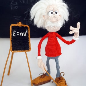 May include: A crocheted doll of a scientist with wild white hair, a red long-sleeved shirt, and gray trousers. The doll stands next to a small black chalkboard on a wooden easel with the white text E=mc2 written on it. The doll holds a pointer.