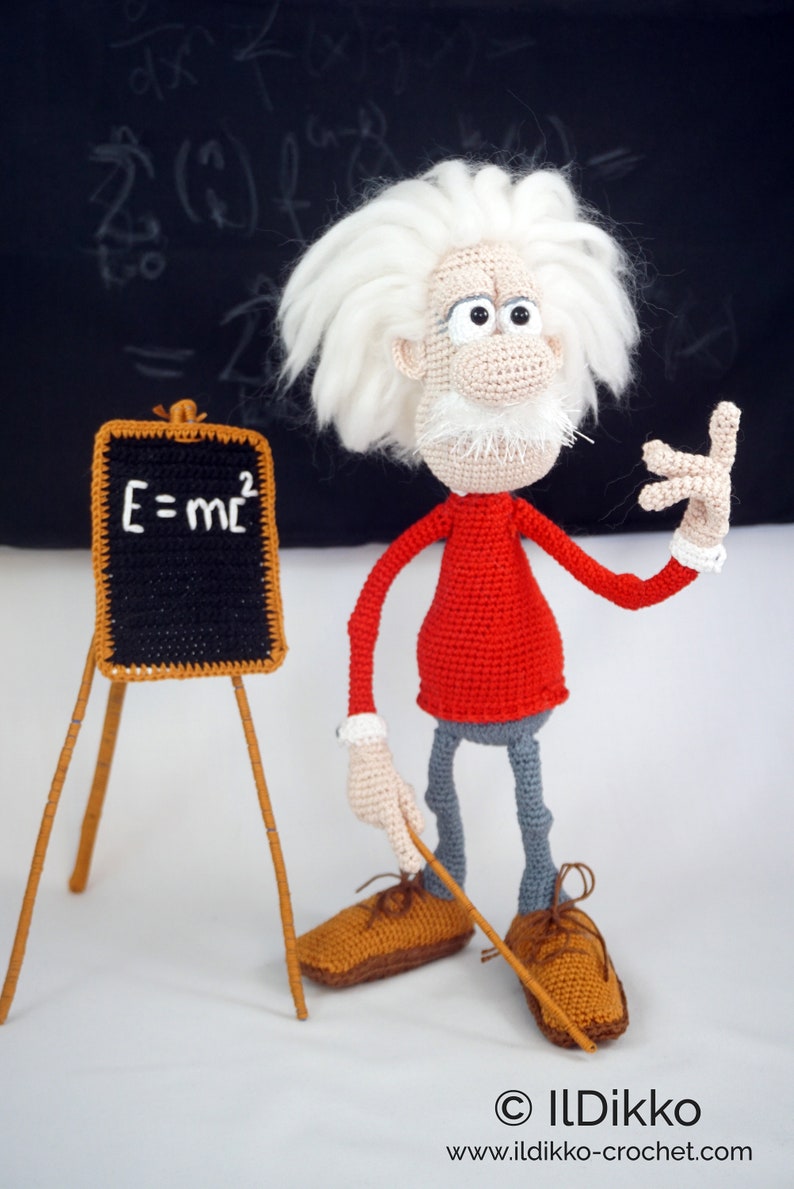 May include: A crocheted doll of a scientist with wild white hair, a red long-sleeved shirt, and gray trousers. The doll stands next to a small black chalkboard on a wooden easel with the white text E=mc2 written on it. The doll holds a pointer.