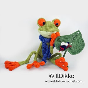 May include: A green crocheted frog wearing a blue scarf sits with a green crocheted leaf. The frog has orange feet and large orange eyes. A small black and white crocheted bug sits on the leaf.