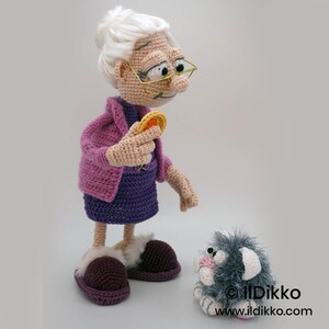 May include: Crocheted doll of an elderly woman wearing a purple dress, pink jacket, and slippers. She holds a yellow object and is looking at a gray cat doll with pink accents. The dolls are handmade.