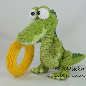 May include: A green crocheted alligator toy with large white eyes and a yellow crocheted ring in its mouth. The toy is sitting on a white surface. © Ildikko www.ildikko-crochet.com