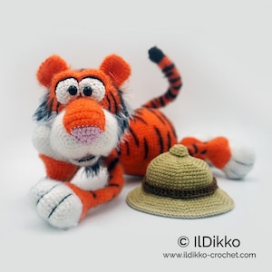 May include: A crocheted orange and black tiger plush toy with white accents, lying down with its head turned to the side. The tiger is wearing a crocheted brown and beige hat.