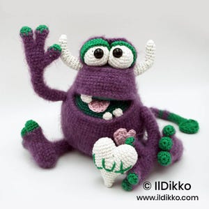 Puede incluir: Un juguete de monstruo tejido a ganchillo de color morado con detalles verdes en dedos, pies y cola. Tiene dos cuernos blancos, grandes ojos negros y una boca ancha que muestra dientes y una lengua rosa. Sostiene un coraz&oacute;n blanco. Texto: IlDikko.