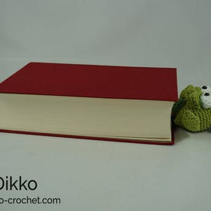 May include: A green crocheted alligator bookmark peeking out from behind a red hardcover book.