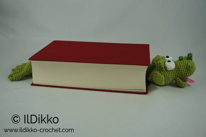 May include: A green crocheted alligator bookmark peeking out from behind a red hardcover book.