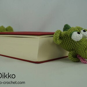 May include: A green crocheted dragon bookmark with large white eyes and a pink tongue sticking out. The bookmark is on top of a closed book with a red cover.
