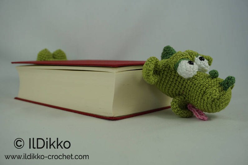 May include: A green crocheted dragon bookmark with large white eyes and a pink tongue sticking out. The bookmark is on top of a closed book with a red cover.