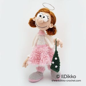 May include: A crocheted doll with auburn hair, a silver wire halo, and a light gray top. The doll wears a fluffy pink skirt, light gray boots with pink trim, and holds a small dark green crocheted tree ornament with white beads. Text: ILDikko
