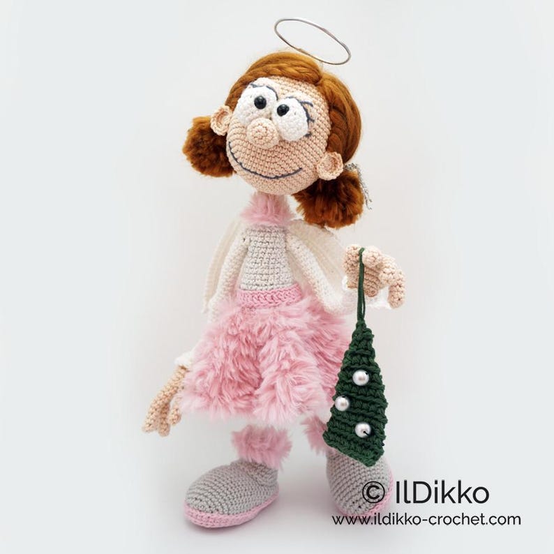 May include: A crocheted doll with auburn hair, a silver wire halo, and a light gray top. The doll wears a fluffy pink skirt, light gray boots with pink trim, and holds a small dark green crocheted tree ornament with white beads. Text: ILDikko