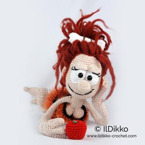 May include: A crocheted doll with messy red yarn hair, a white bone accessory, and large white eyes. The doll has a beige complexion, wears an orange and black patterned garment, and rests one hand on its cheek next to a small red crocheted apple.