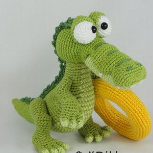 May include: A green crocheted crocodile toy with large white eyes and a yellow crocheted ring held in its mouth. The toy features a textured back and webbed feet. The text "&copy; ILDikko www.ildikko-crochet.com" is printed at the bottom.