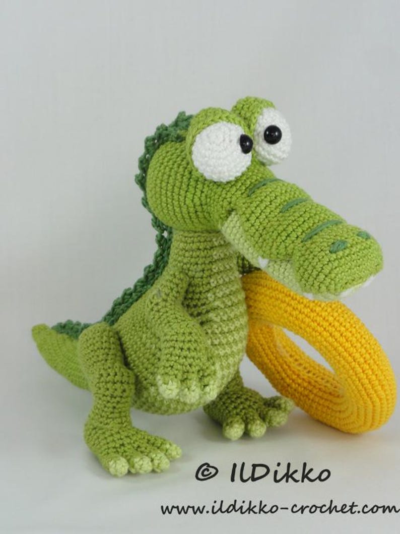 May include: A green crocheted crocodile toy with large white eyes and a yellow crocheted ring held in its mouth. The toy features a textured back and webbed feet. The text "&copy; ILDikko www.ildikko-crochet.com" is printed at the bottom.