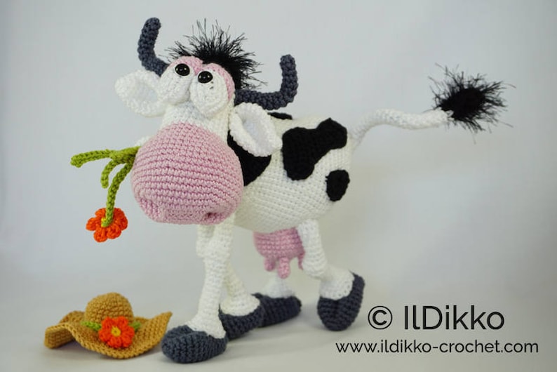 May include: A crocheted cow toy with black and white spots, pink ears, and a black tail. The cow is holding a green stem with an orange flower. The cow is standing on a white surface with a brown crocheted hat with an orange flower on it.
