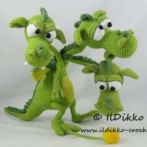 May include: Three green crocheted dragons with large eyes and toothy grins. The dragons are standing on a white surface with a yellow flower in the foreground. The text "© IlDikko www.ildikko-crochet.com" is visible in the bottom right corner of the image.