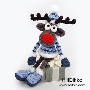 May include: A crocheted reindeer toy with a bright red nose, dark brown antlers, and large white eyes. It wears a blue and white striped sweater, matching hat with a white pom-pom, and blue boots with white trim. It sits atop a gray gift box.