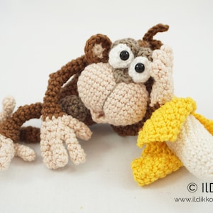 Puede incluir: Un juguete de mono de crochet con pelaje marrón, manos y pies blancos y grandes ojos negros. El mono sostiene un plátano de crochet con una cáscara amarilla y un cuerpo blanco.