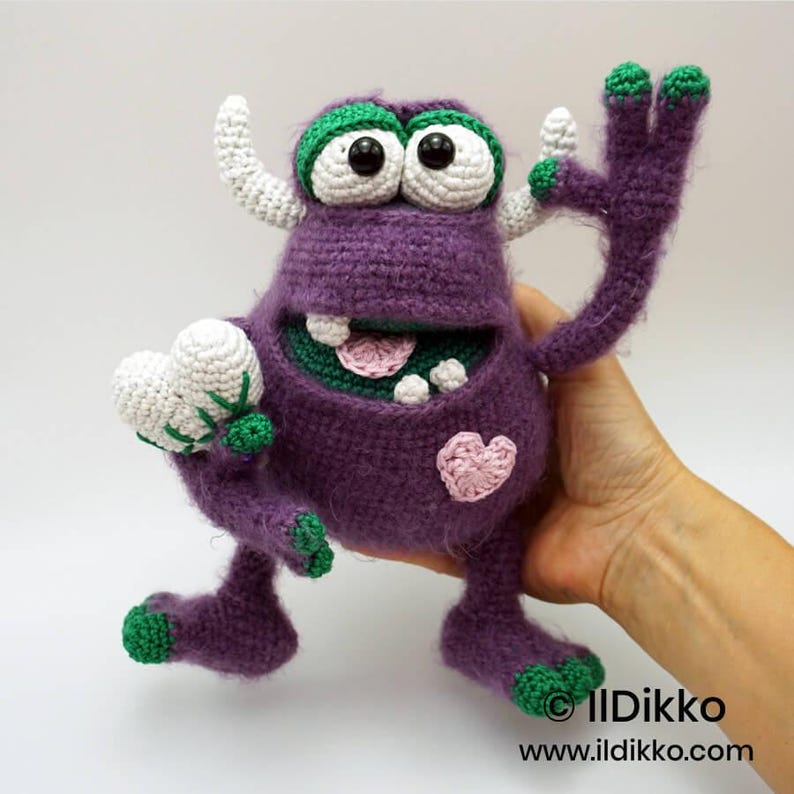 Puede incluir: Un juguete de monstruo tejido a ganchillo en color morado con cuernos blancos, ojos grandes blancos con pupilas negras y detalles verdes en pies y manos. Tiene una boca ancha con dientes blancos, lengua rosa y un peque&ntilde;o parche de coraz&oacute;n rosa.