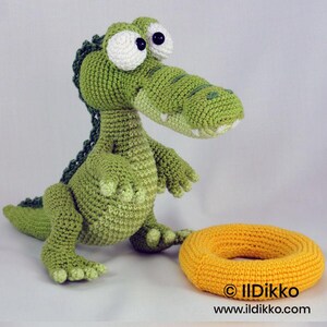 May include: A green crocheted crocodile toy with large white eyes and black pupils, featuring a textured back ridge and a light green belly. Beside the crocodile sits a round, yellow crocheted ring. The text "&copy; Ildikko www.ildikko.com" is visible.