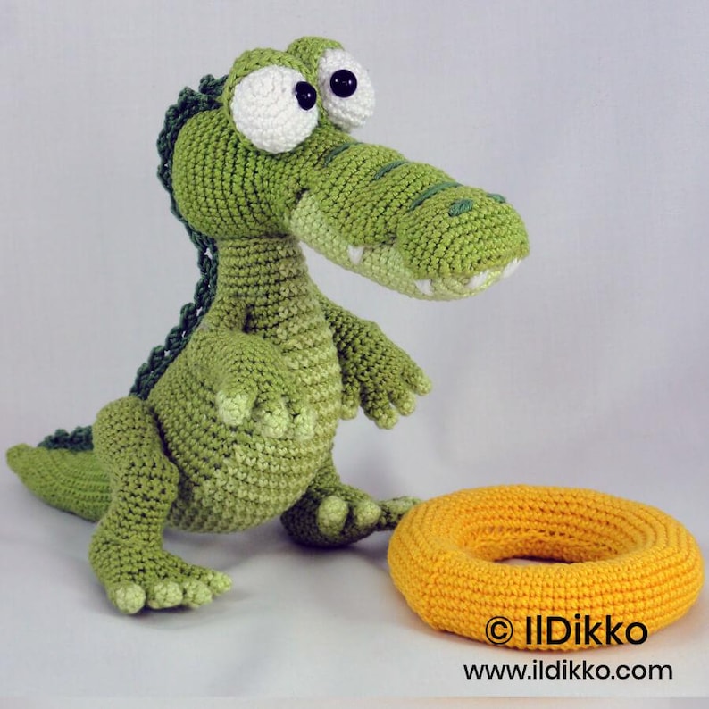 May include: A green crocheted crocodile toy with large white eyes and black pupils, featuring a textured back ridge and a light green belly. Beside the crocodile sits a round, yellow crocheted ring. The text "&copy; Ildikko www.ildikko.com" is visible.