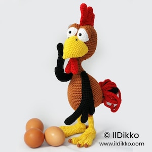 Puede incluir: Un gallo de ganchillo hecho a mano con cresta, barbilla y cola rojas, pico y patas amarillas y grandes ojos con bordes blancos. El gallo es marrón, negro y amarillo. Tres huevos marrones están en la base del gallo. Incluye el texto "© Ildikko" y "www.ildikko.com".