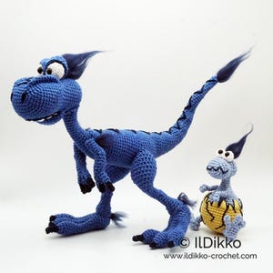 May include: A large blue crocheted dinosaur with dark blue tufts of hair and black claws, standing next to a smaller light blue crocheted dinosaur emerging from a yellow cracked egg. The text ILDikko and www.ildikko-crochet.com is visible.