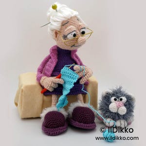 May include: A handmade crochet doll of a grandmother knitting with a blue yarn. The doll wears a purple dress, pink jacket, and purple shoes with white fur trim. A gray cat doll sits beside her. The doll has glasses and white hair.