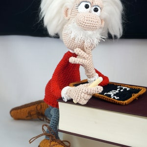 May include: A crocheted doll of a scientist with wild white hair, a white beard, and a red shirt, sitting on a large book. The doll holds a small piece of chalk and rests a hand on a miniature blackboard with the text E=mc2 written in white.