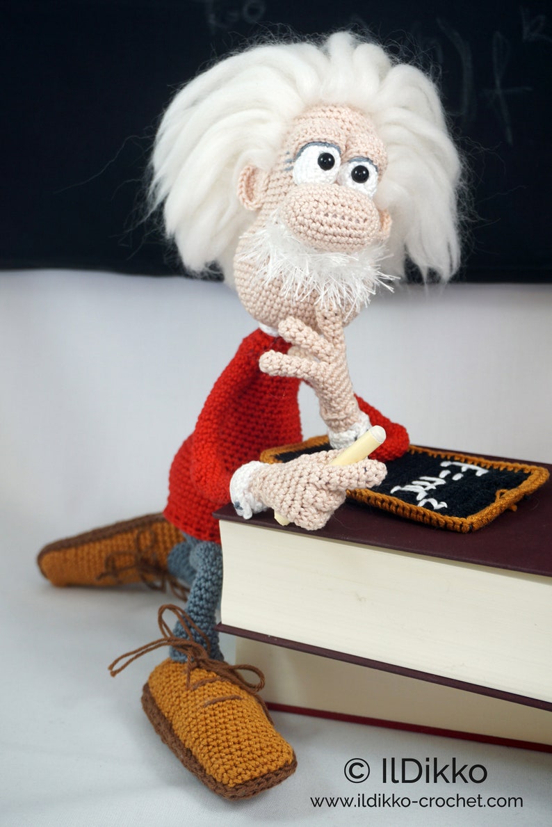 May include: A crocheted doll of a scientist with wild white hair, a white beard, and a red shirt, sitting on a large book. The doll holds a small piece of chalk and rests a hand on a miniature blackboard with the text E=mc2 written in white.