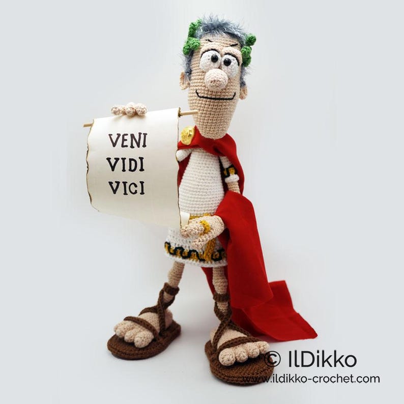 以下が含まれることがあります： 「veni vidi vici」というラテン語のフレーズが書かれた巻物を手に持つ、ローマ兵士の編みぐるみフィギュア。兵士は白いチュニック、赤いマント、月桂樹の冠を着用しています。