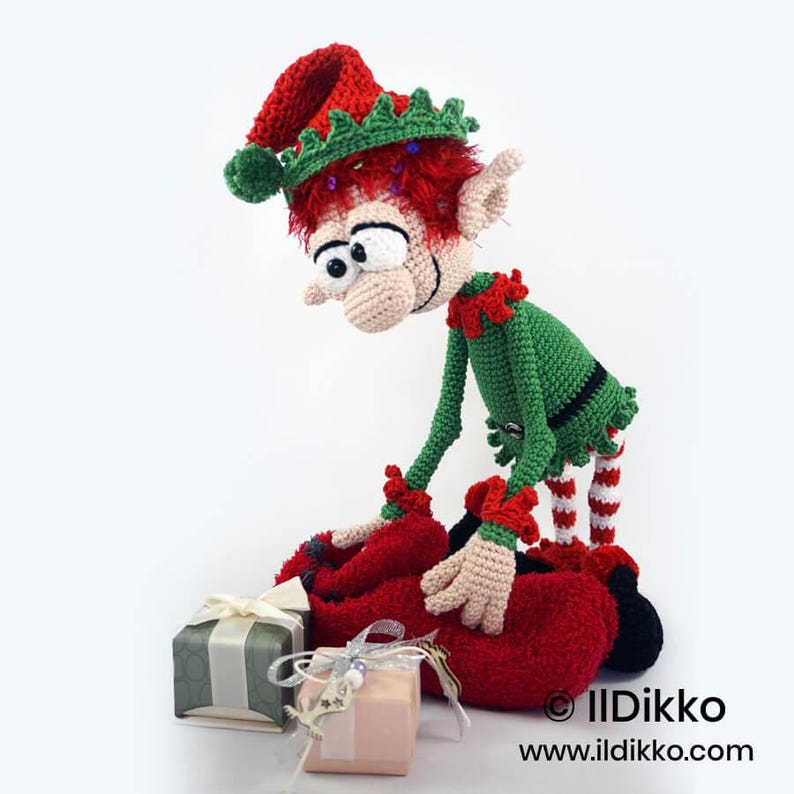 May include: A crocheted elf doll with red hair, a green and red outfit, and a pointed hat, leaning over two small gift boxes wrapped with ribbons. The doll has large eyes and is positioned next to the text IIDikko www.ildikko.com.