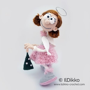 May include: A crocheted doll with brown hair, a silver wire halo, and a pink fuzzy skirt. The doll has white wings, grey shoes, and holds a small dark green triangular ornament decorated with white beads. Text reads ILDikko ildikko-crochet.com.