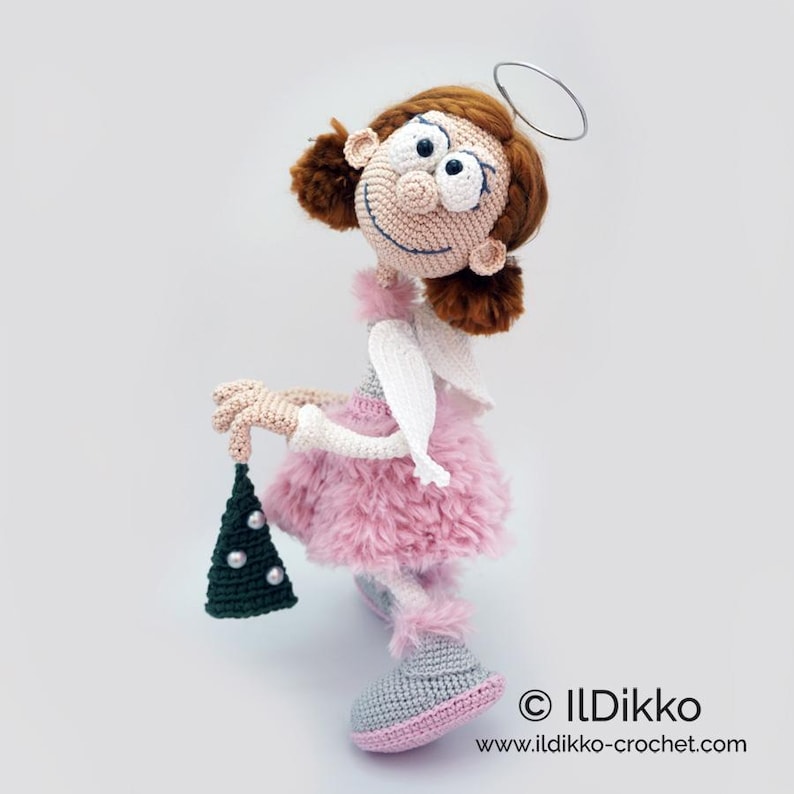 May include: A crocheted doll with brown hair, a silver wire halo, and a pink fuzzy skirt. The doll has white wings, grey shoes, and holds a small dark green triangular ornament decorated with white beads. Text reads ILDikko ildikko-crochet.com.