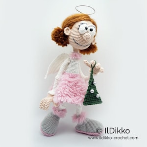 May include: A crocheted doll with auburn hair, a silver halo, and white wings. The doll wears a pink fuzzy skirt, white top, and gray boots with pink trim. It holds a small green crocheted Christmas tree with silver beads. Text reads ILDikko.