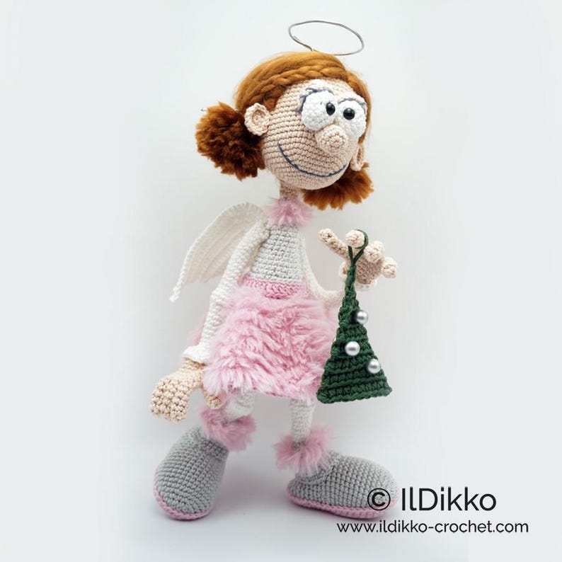 May include: A crocheted doll with auburn hair, a silver halo, and white wings. The doll wears a pink fuzzy skirt, white top, and gray boots with pink trim. It holds a small green crocheted Christmas tree with silver beads. Text reads ILDikko.