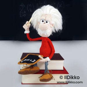 May include: A crocheted doll of an elderly person with wild white hair, a white mustache, and a red long-sleeved shirt. The doll sits on two stacked books, holding a small black chalkboard with the white text E=mc2 and a small wooden pointer.