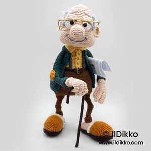 May include: A crocheted doll of an elderly man wearing glasses, a green jacket, and brown pants. He holds a cane and a newspaper. The doll has a smiling face and is wearing brown and tan slippers.