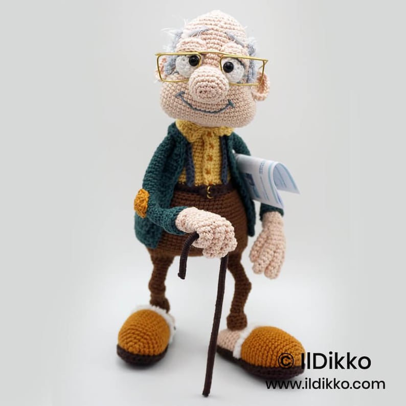 May include: A crocheted doll of an elderly man wearing glasses, a green jacket, and brown pants. He holds a cane and a newspaper. The doll has a smiling face and is wearing brown and tan slippers.