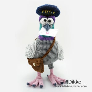 May include: A crocheted pigeon wearing a blue hat with the word "POST" on it, holding a white envelope with a pink heart. The pigeon has a brown bag slung over its shoulder and pink feet.