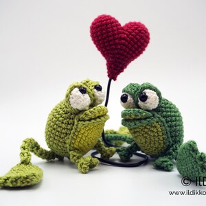 May include: Two green crocheted frogs with large eyes and a red crocheted heart balloon. The frogs are facing each other and the balloon is between them.