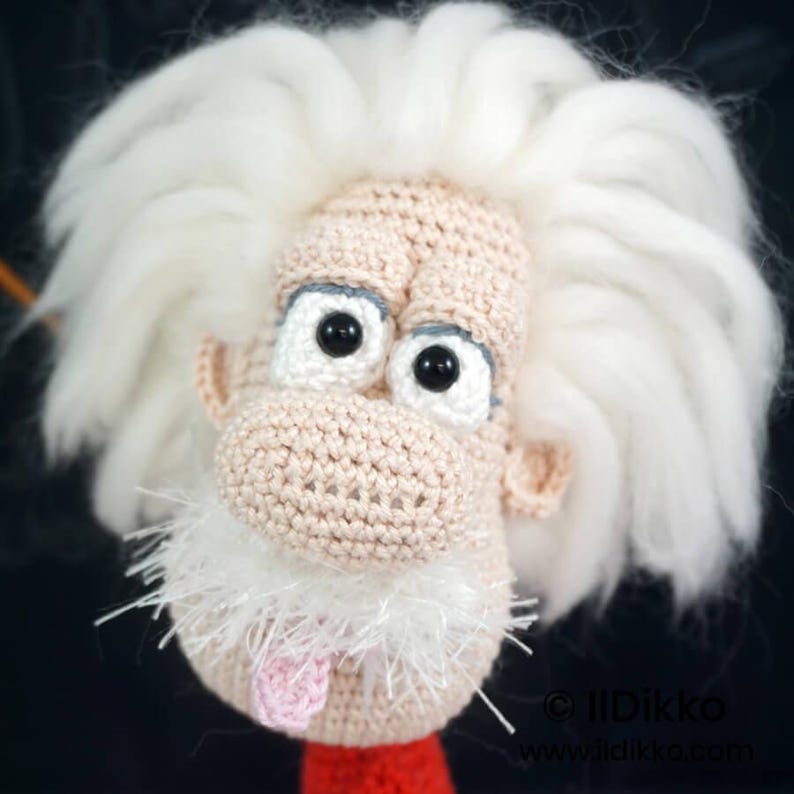 May include: A crocheted doll head featuring a light beige complexion, large black plastic eyes, and a prominent white yarn hairstyle. The figure has a textured white mustache and a small pink tongue protruding from the mouth area. Text reads IIDikko.